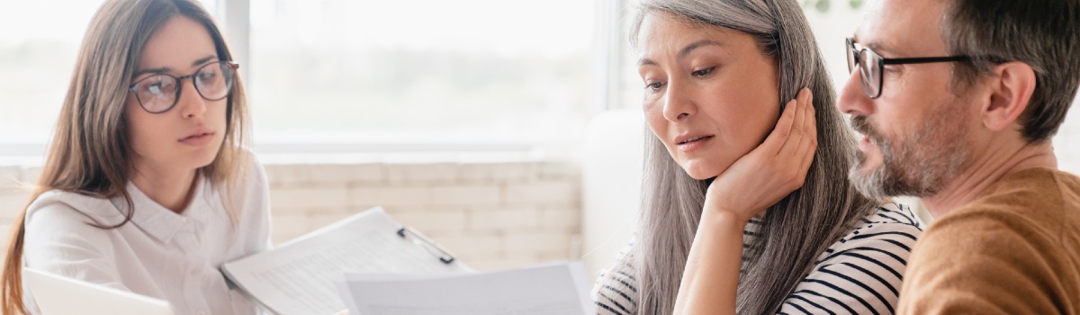 A middle-aged couple looks over documents with concern as a young professional woman points at information on a page to them.